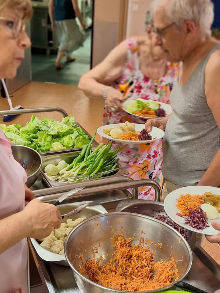 Des seniors se servent des plats lors d’un repas collectif, prenant des légumes et des salades sur un chariot de service.